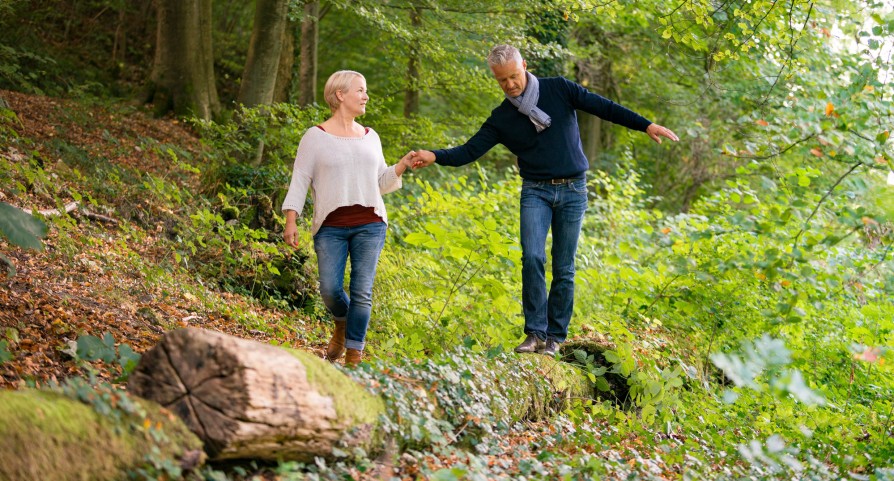 Frau hält Mann, der über einen Baumstamm balanciert, die Hand. Une femme donnant la main à un homme en équilibre sur un tronc d'arbre. Una donna tiene la mano a un uomo in equilibrio su un tronco d'albero.