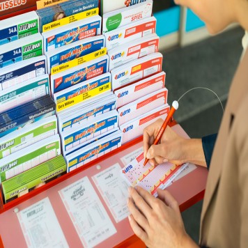 Ein Frau steht an einem Spielscheinständer und füllt einen Swiss Lotto Spielschein aus.